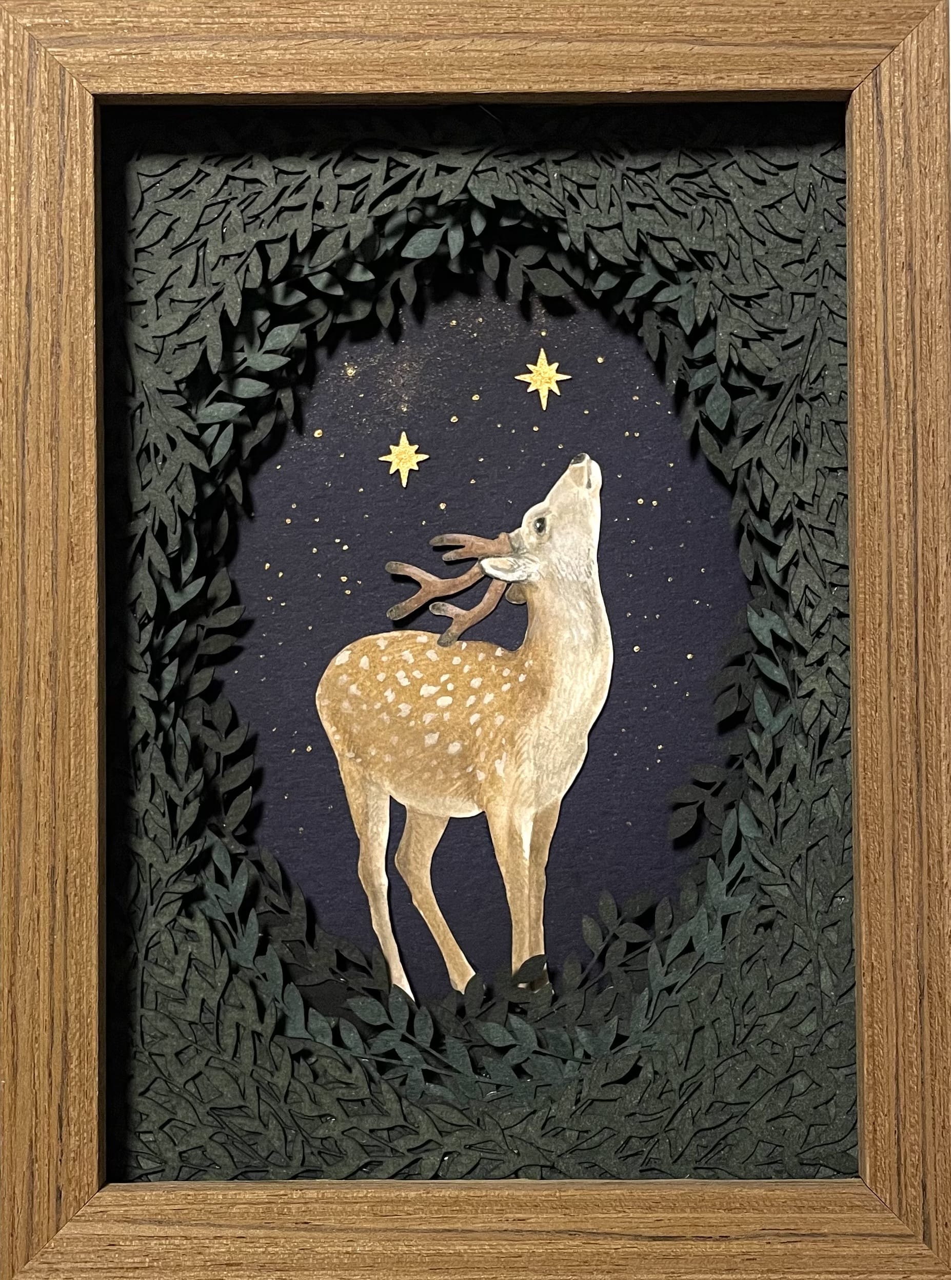 a small paper collage in a wooden frame of a deer with small glowing stars peeking out from behind a surround of foliage