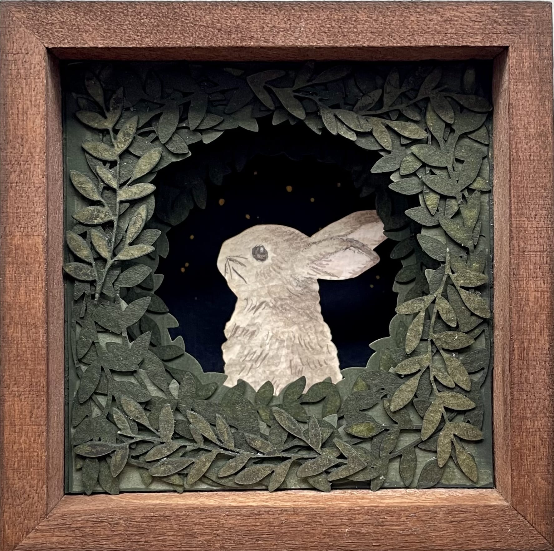 a small paper collage in a wooden frame of a bunny peeking out from behind a surround of foliage
