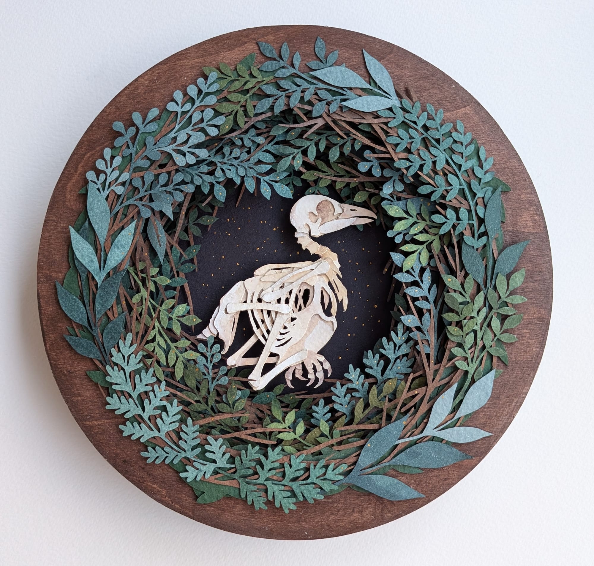 a paper collage on a round surface featuring a wreath-like arrangement of foliage around the skeleton of a bird