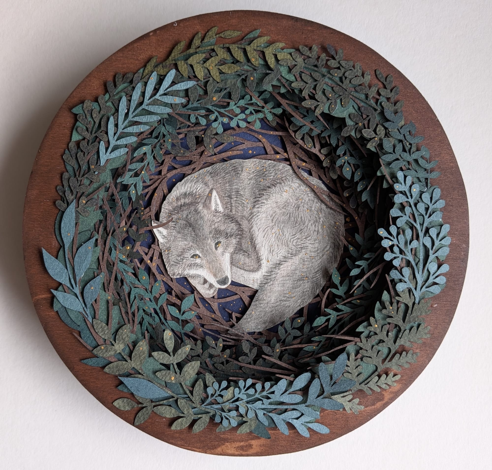 a paper collage on a round surface featuring a wreath-like arrangement of foliage around a large sleeping wolf