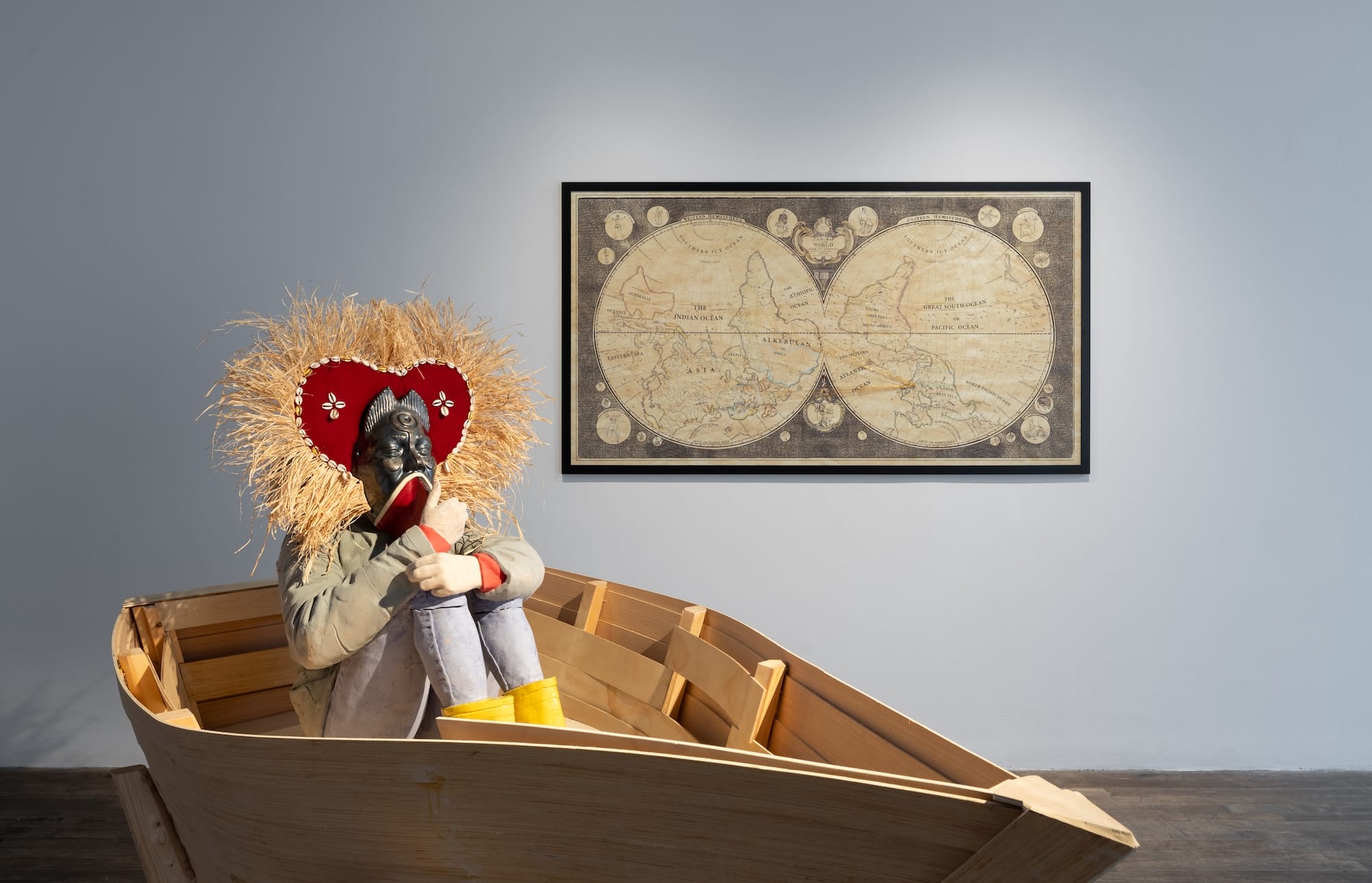 a sculpture of a masked figure with an ornate headdress, sitting in a boat in front of a historic global map