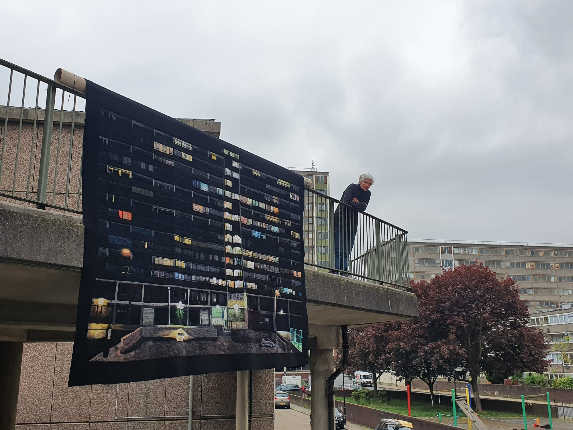 a textile piece depicting public housing hangs over a balcony while the artist leans on the railing nearby