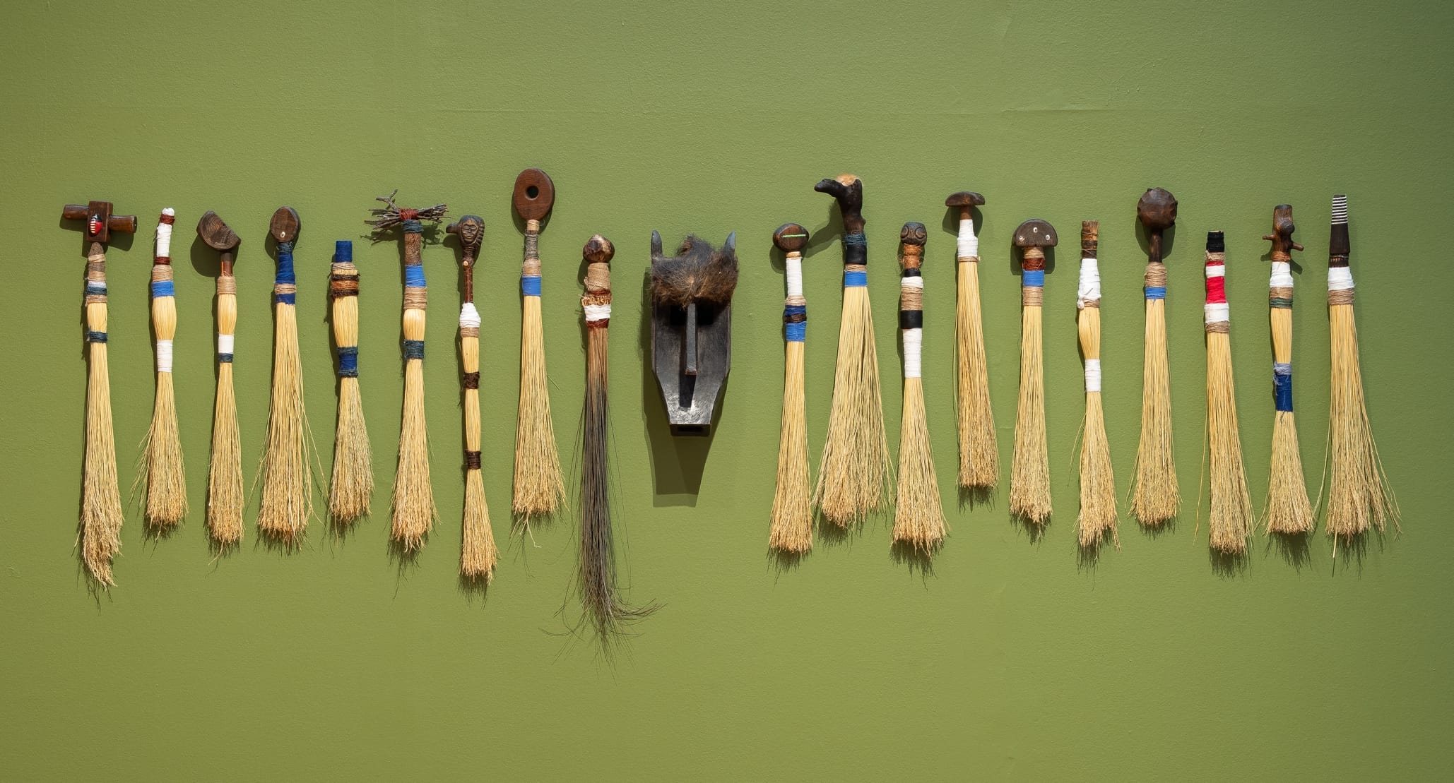 a collection of hand brooms flank an african mask on a green wall
