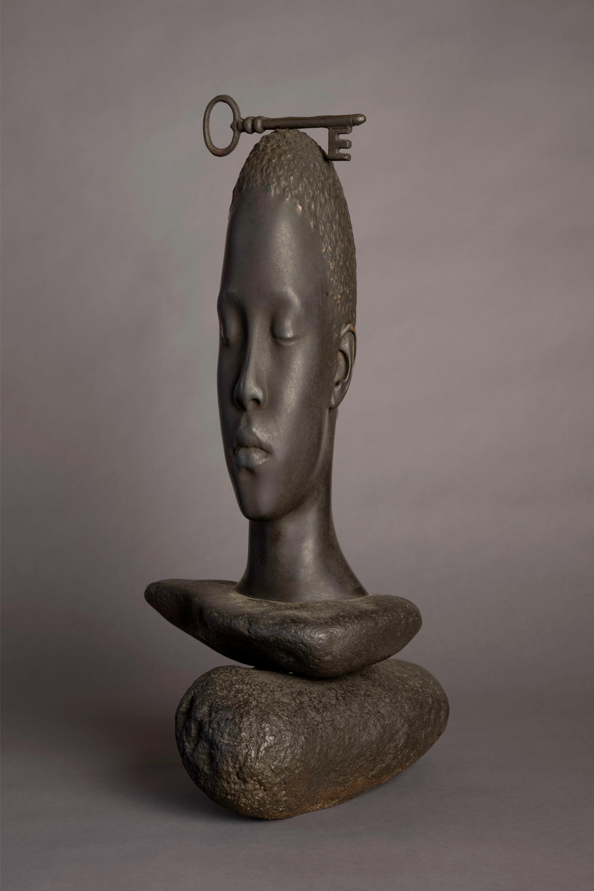 a bronze sculpture by Jaume Plensa of a face resting upon rocks with a large key balancing on its head