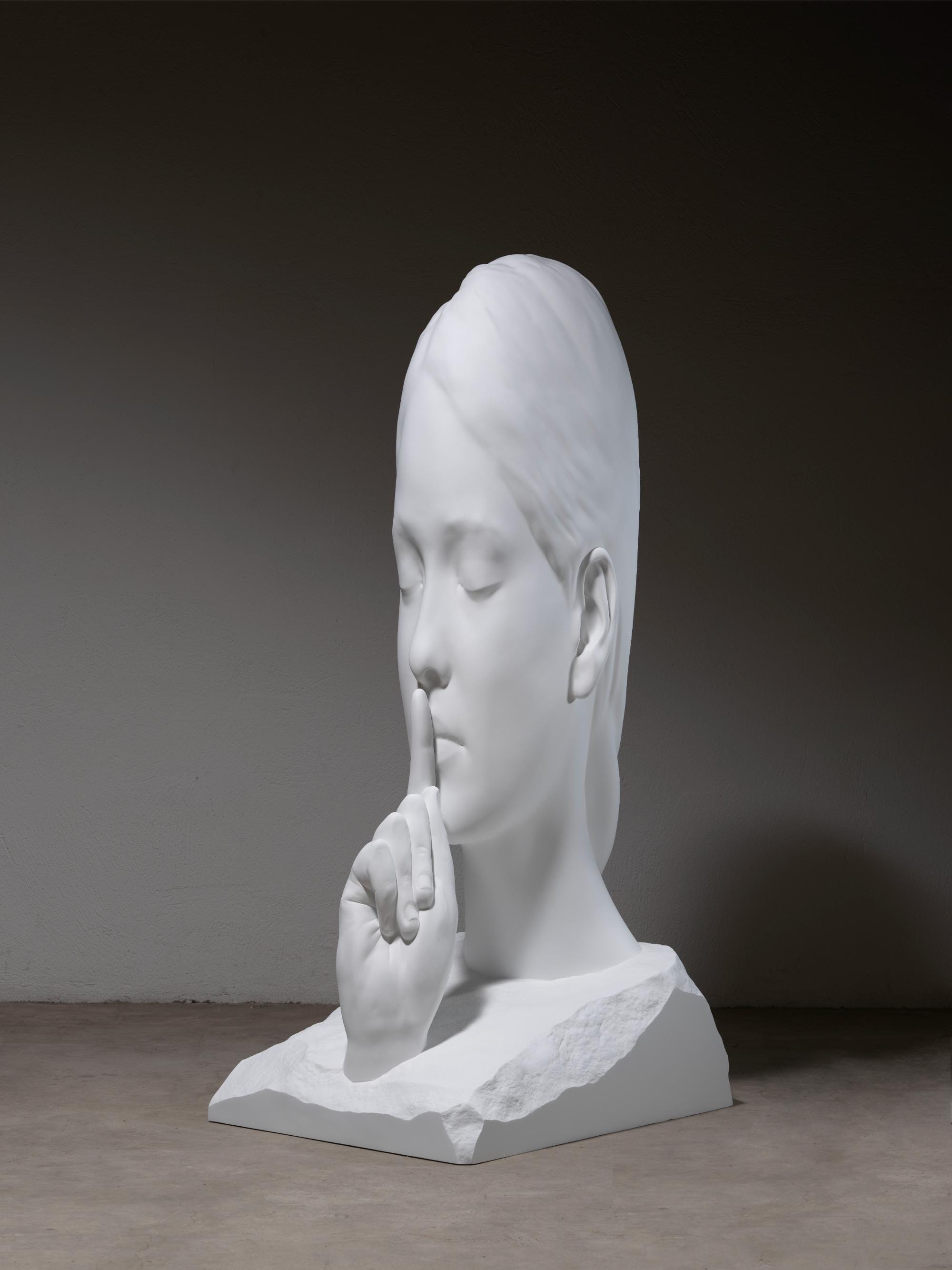 a bronze sculpture by Jaume Plensa of a face with its eyes closed and a finger to its mouth, painted in white