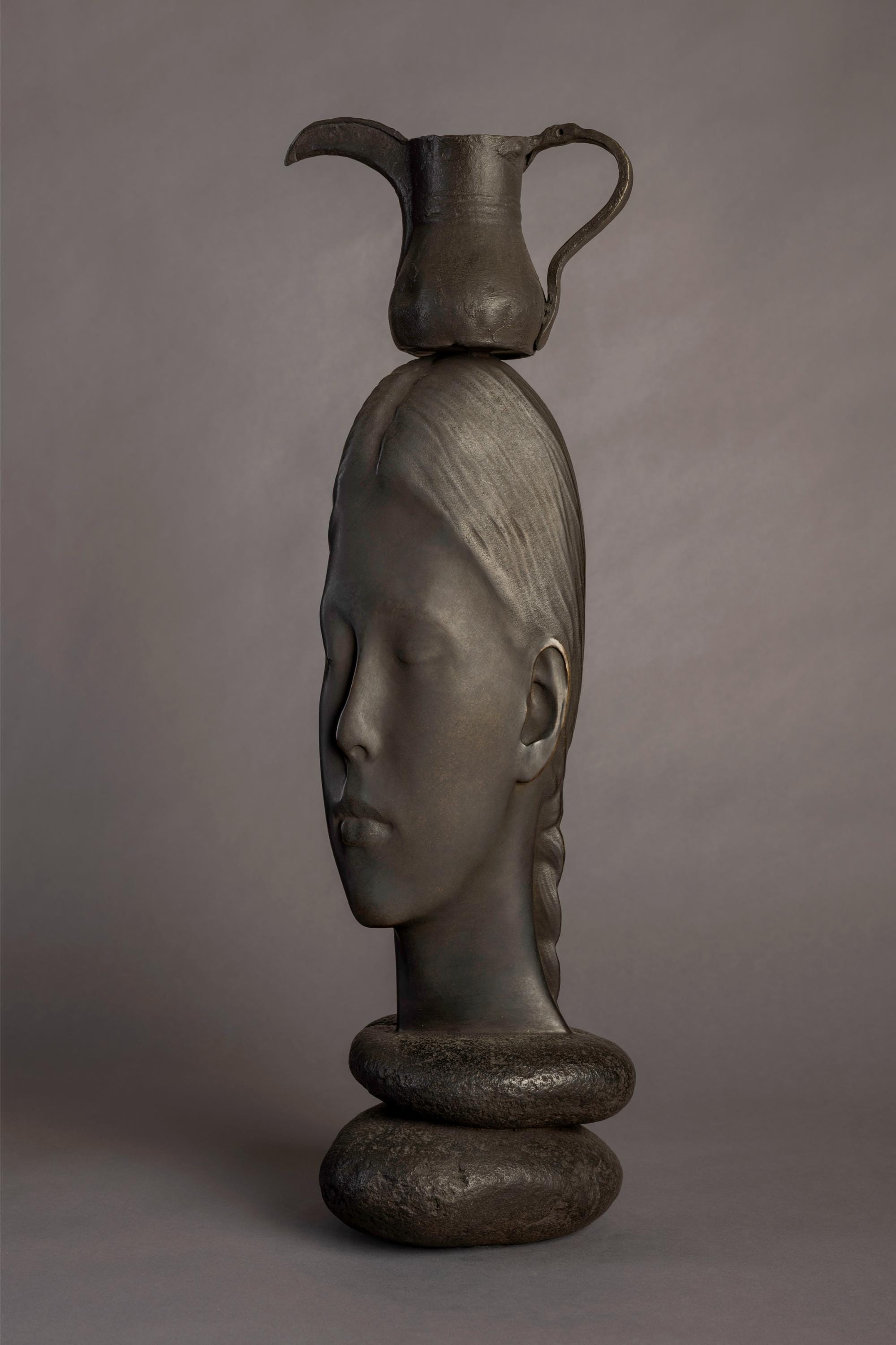 a bronze sculpture by Jaume Plensa of a face resting upon rocks with a large drink jug balancing on its head