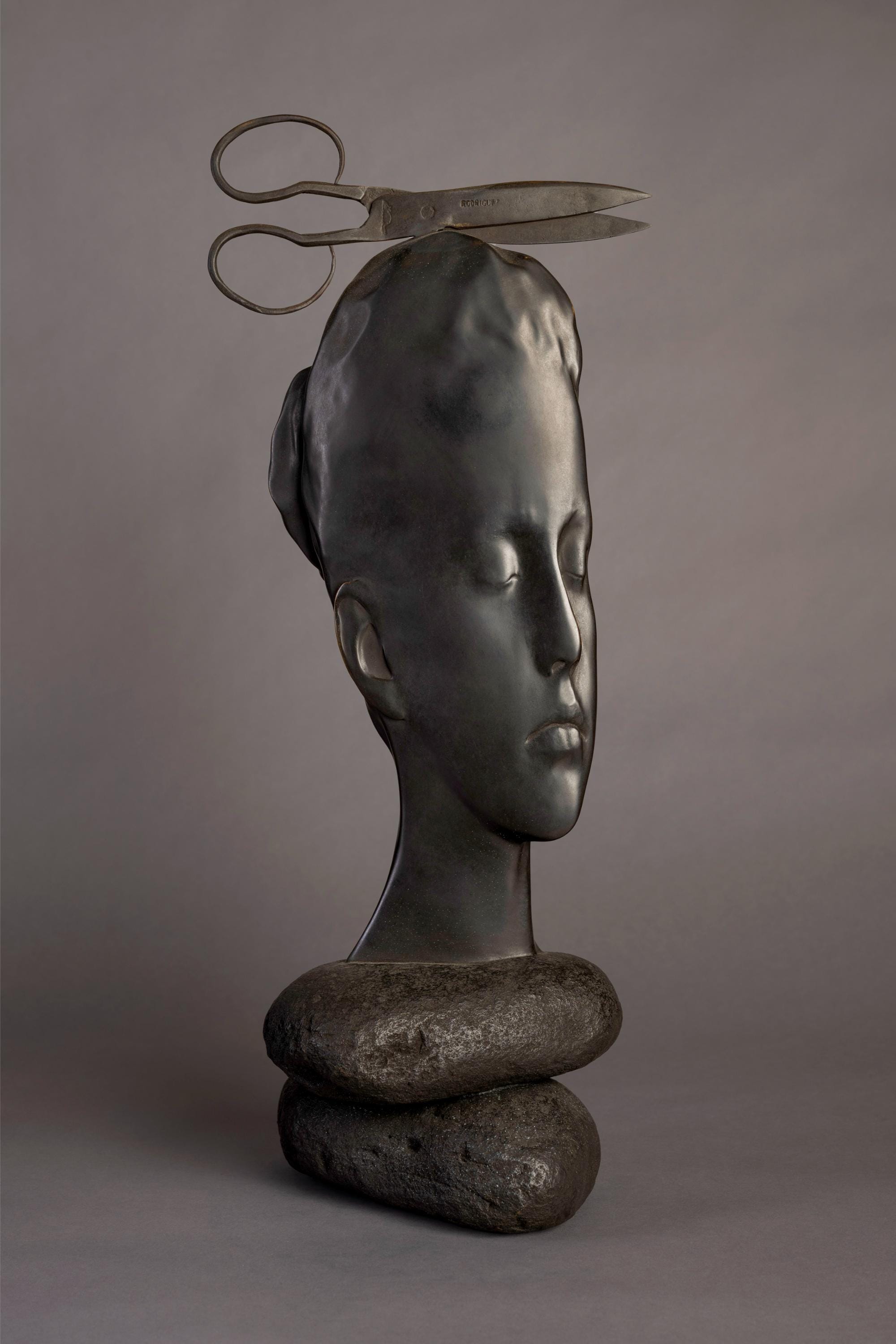 a bronze sculpture by Jaume Plensa of a face resting upon rocks with a large pair of scissors balancing on its head