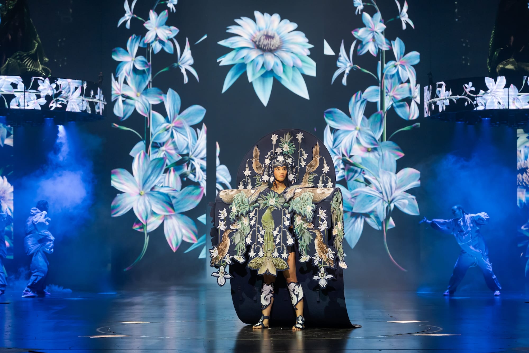 an elaborate handmade garment with blue flowers and a huge cape-like feature, worn by a performer on a stage with projections of blue flowers in the background