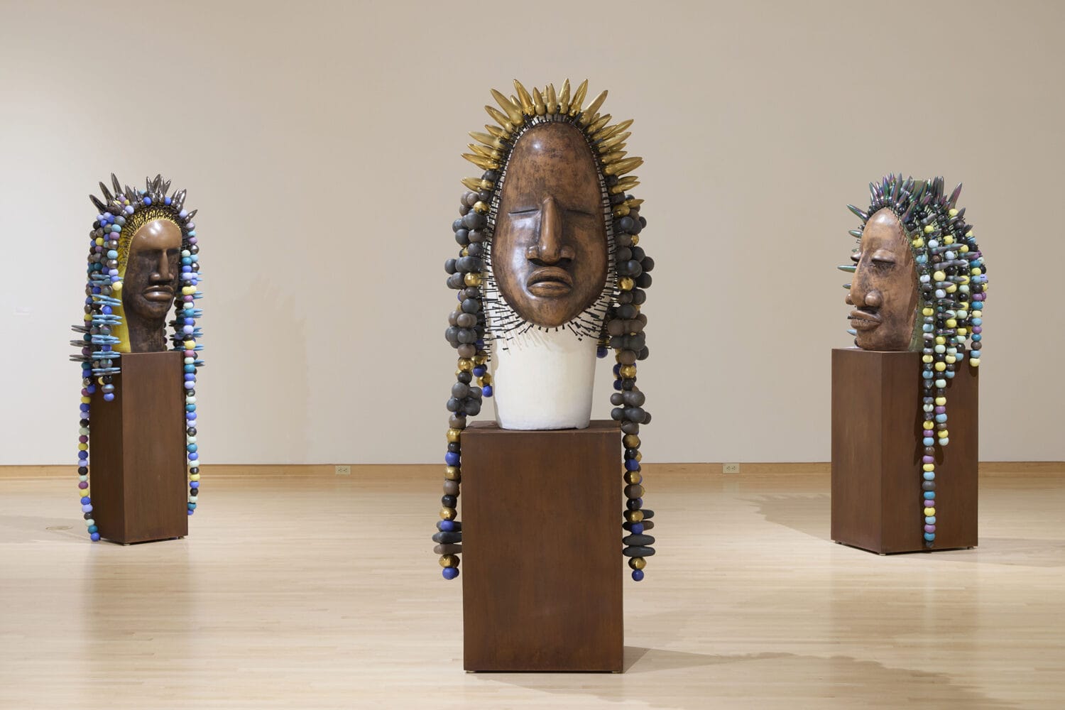 three large sculptures of stylized Black figures' heads, inspired by African masking traditions, with large colorful beads draped like hair or headdresses