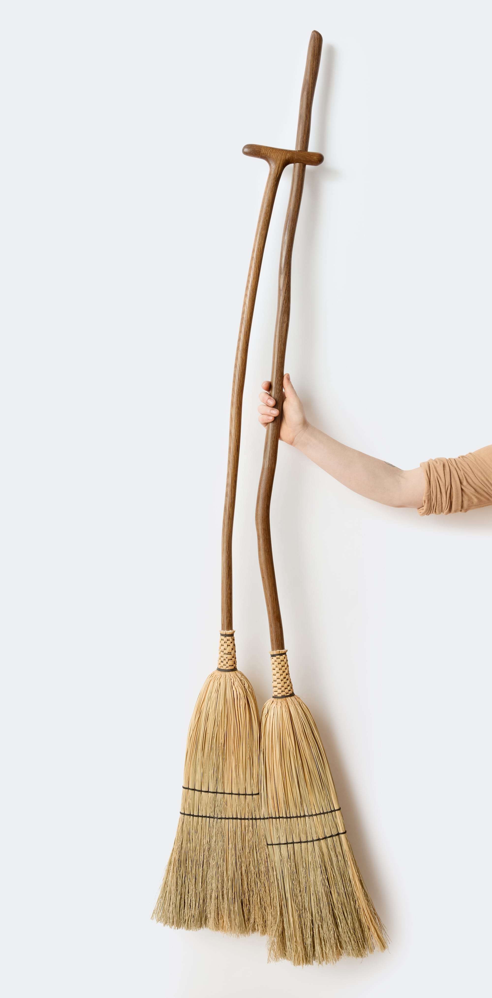 a hand holds a sculptural broom that looks like two conjoined straw brooms in one