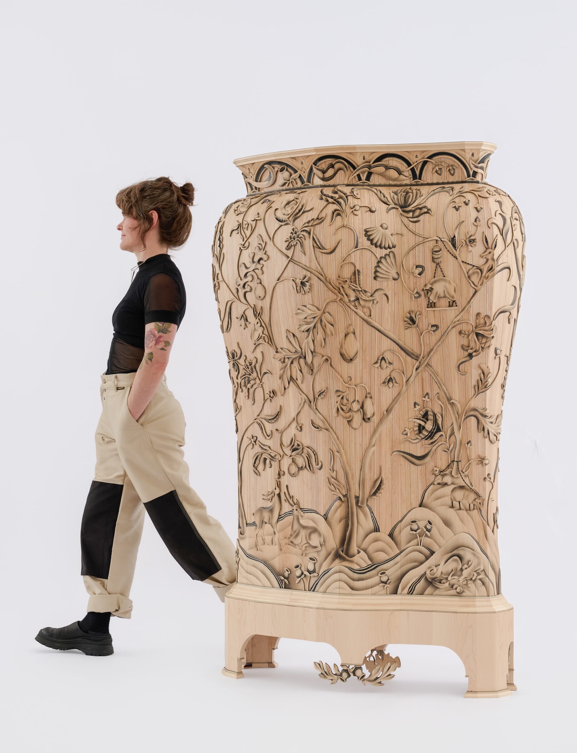 artist Aspen Golann walks by a large wooden cabinet that resembles a giant vessel, with crewel-inspired wood carving on the exterior featuring plants and animals