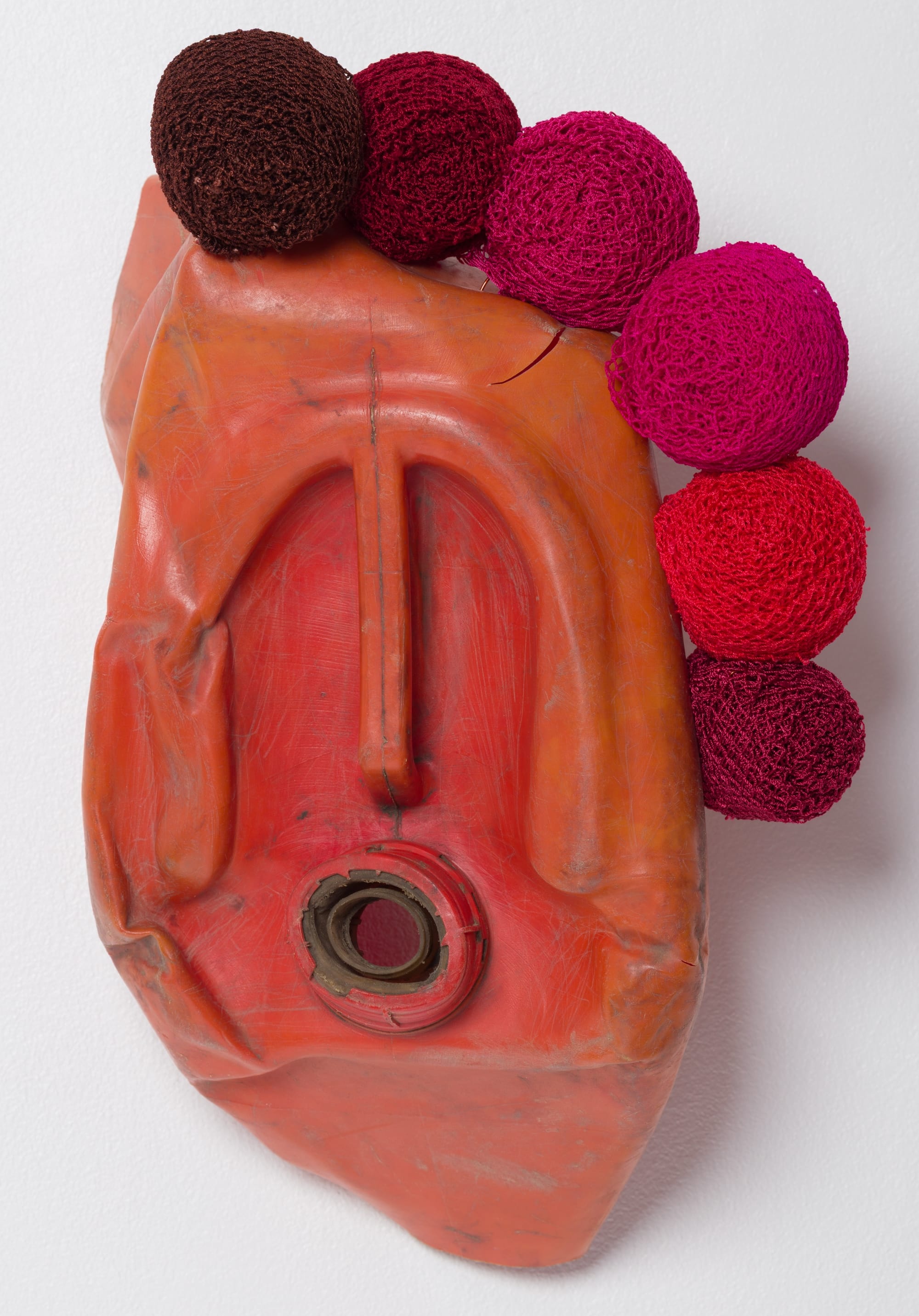 an abstract mask-like sculpture made of an orange plastic jug and red and pink knit spheres