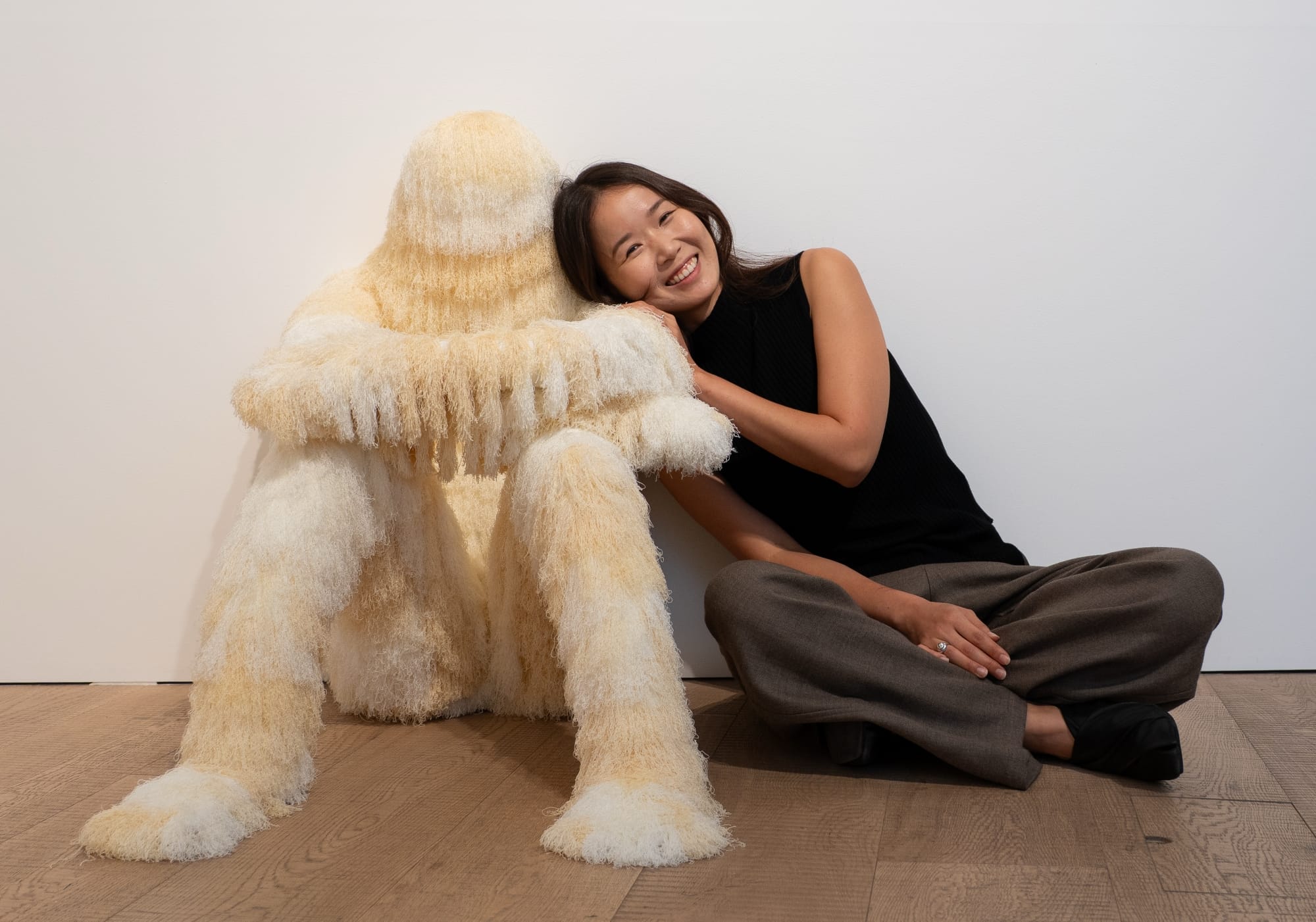 an asian woman in a black top and gray pants sits on the floor to pose with a white and beige fur colored seated figure