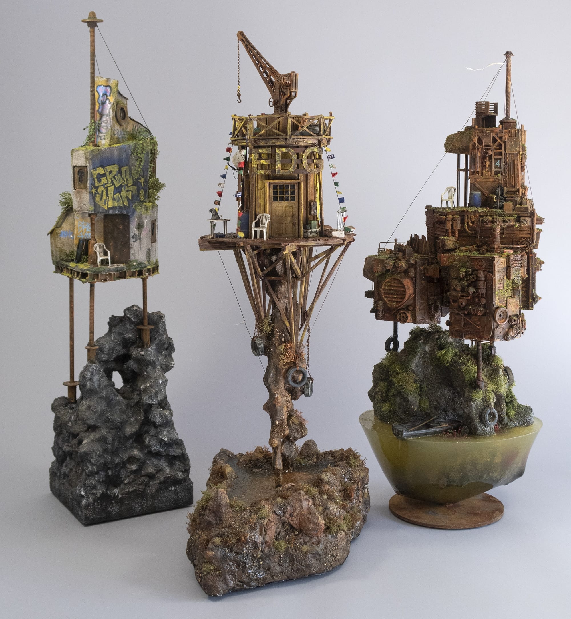 three sculptures by Simon Laveuve in 1/35 scale of ramshackle, dystopian houses built on stilts and rocky outcrops