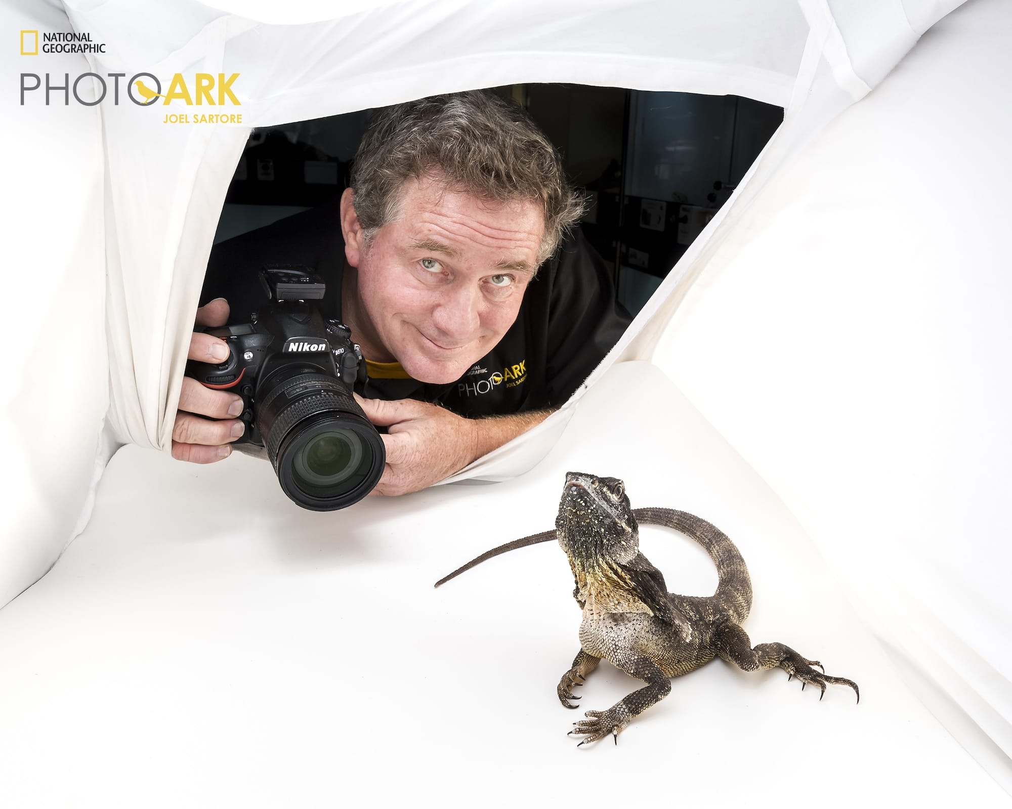 a portrait of National Geographic photographer Joel Sartore holding a camera inside of a white light box with a lizard facing the viewer