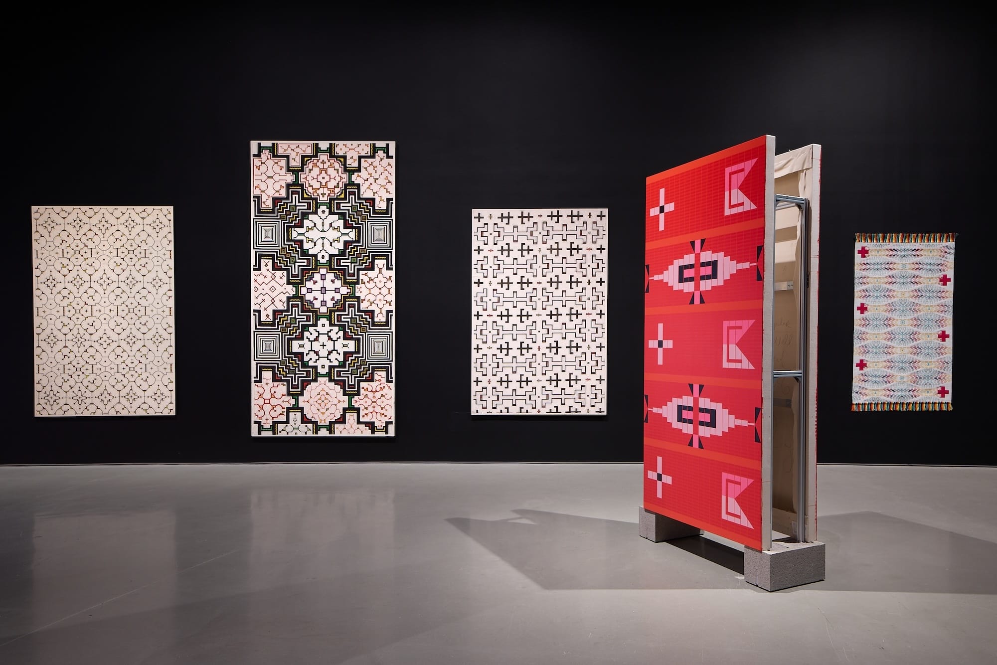 a series of geometric weavings installed in a gallery space on a dark wall