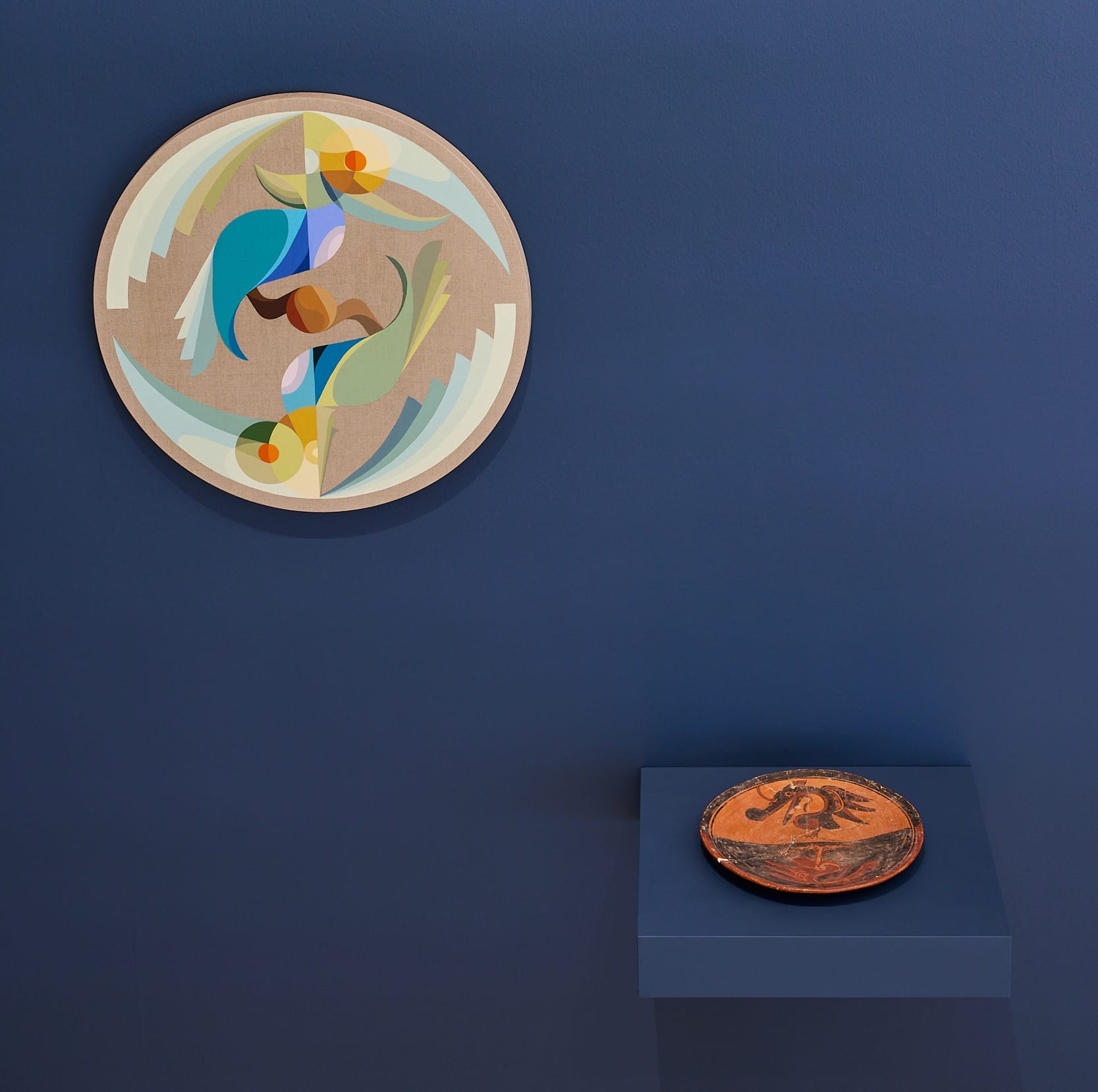 a small tondo by Eamon Ore-Giron featuring a geometric, colorful bird, displayed alongside a historic Mayan ceramic plate with a bird design