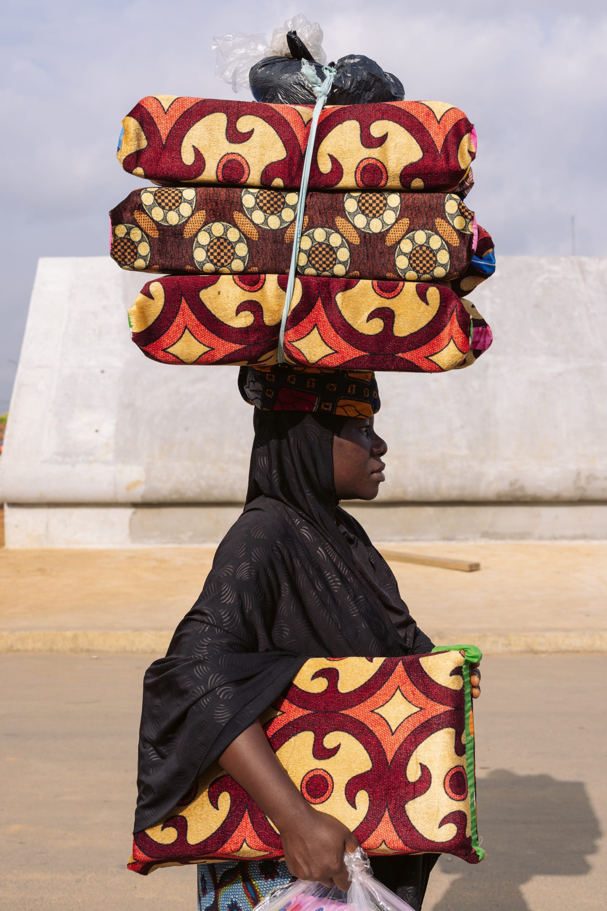 a woman in a black headscarf carries patterned cushions on her head and one under her arm