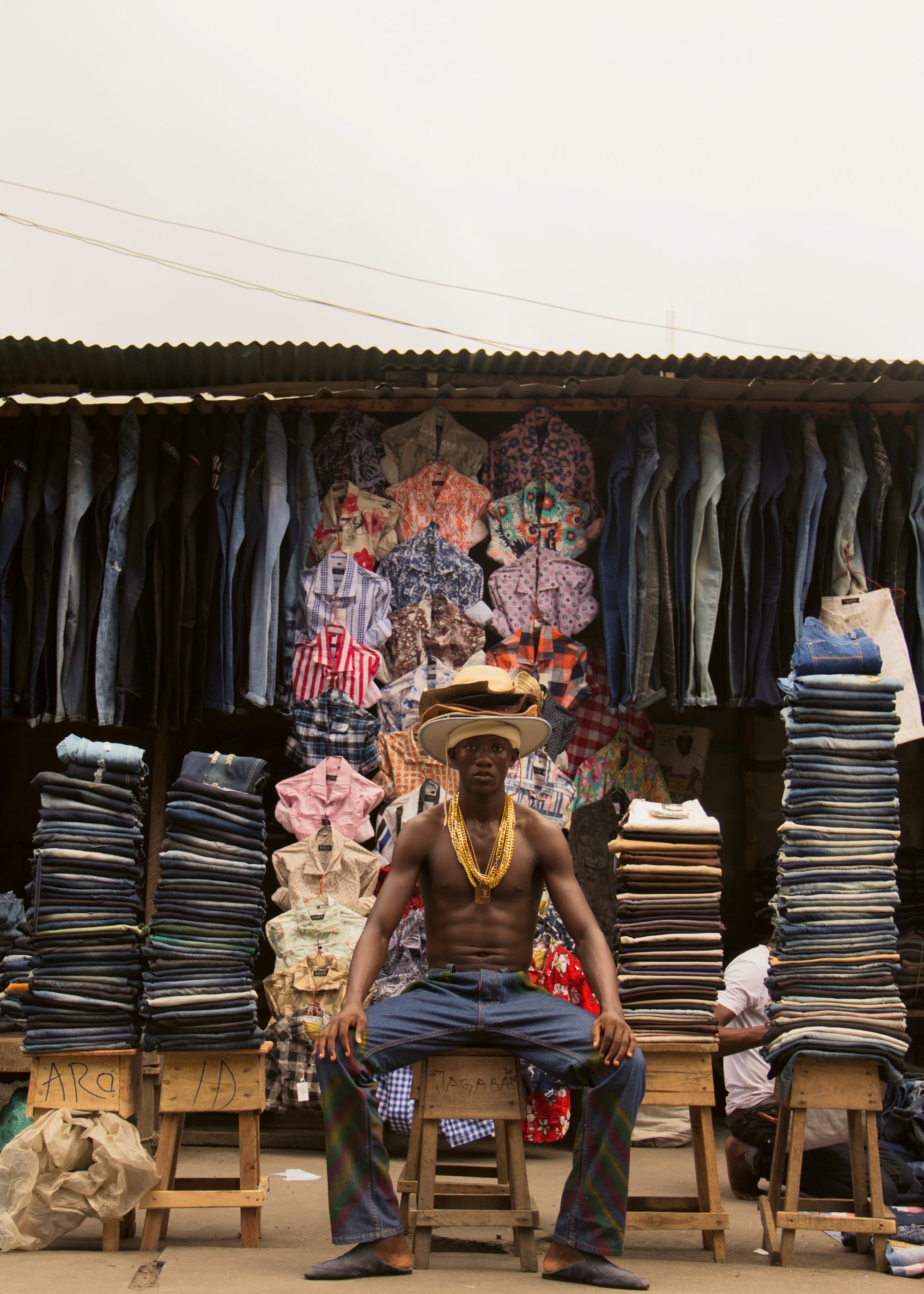 a shirtless man poses while sitting on a wooden stool surrounded by folded and hanging garments