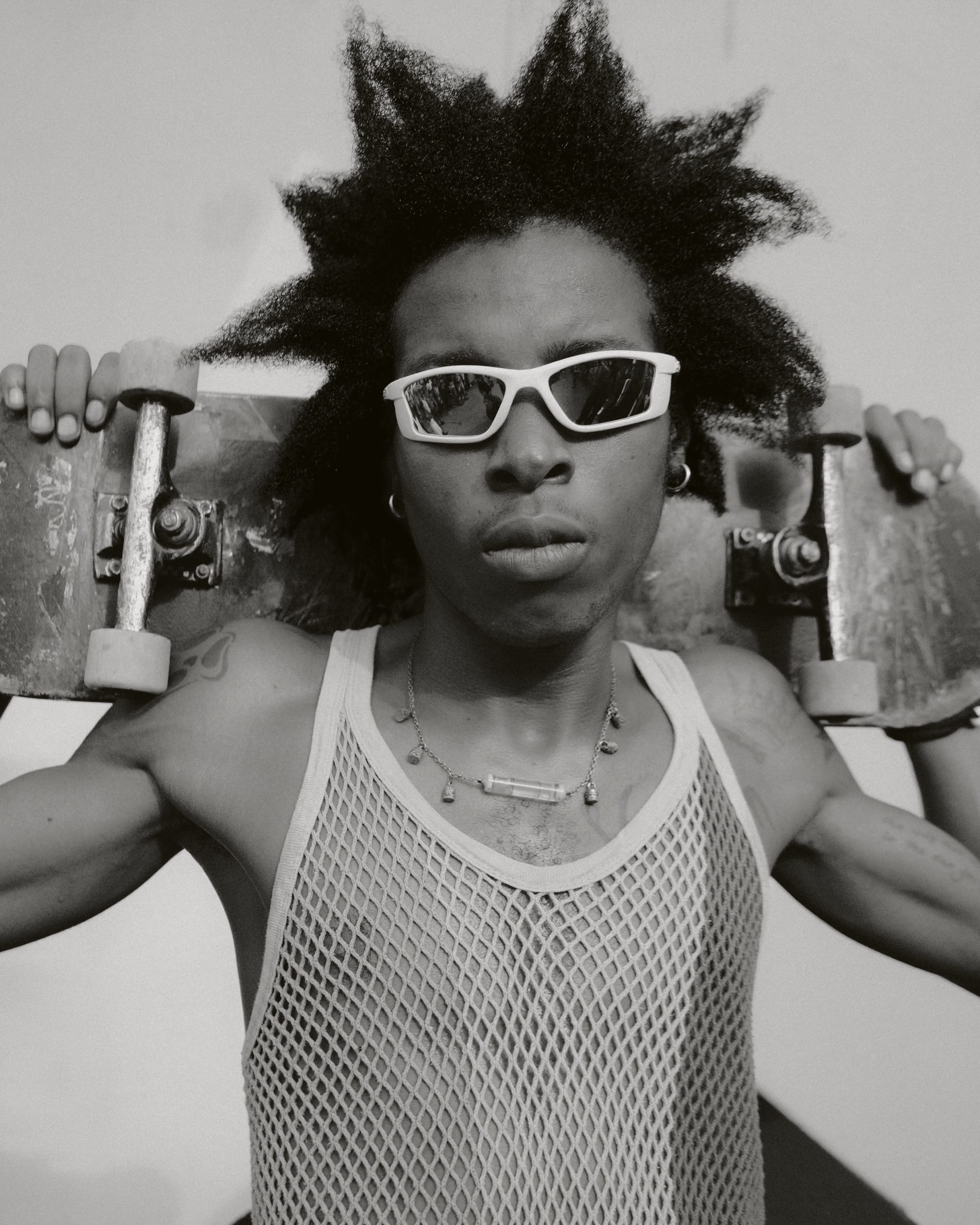 a black and white photo of a person in white rimmed sunglasses and a mesh tank top carrying a skateboard on their shoulders