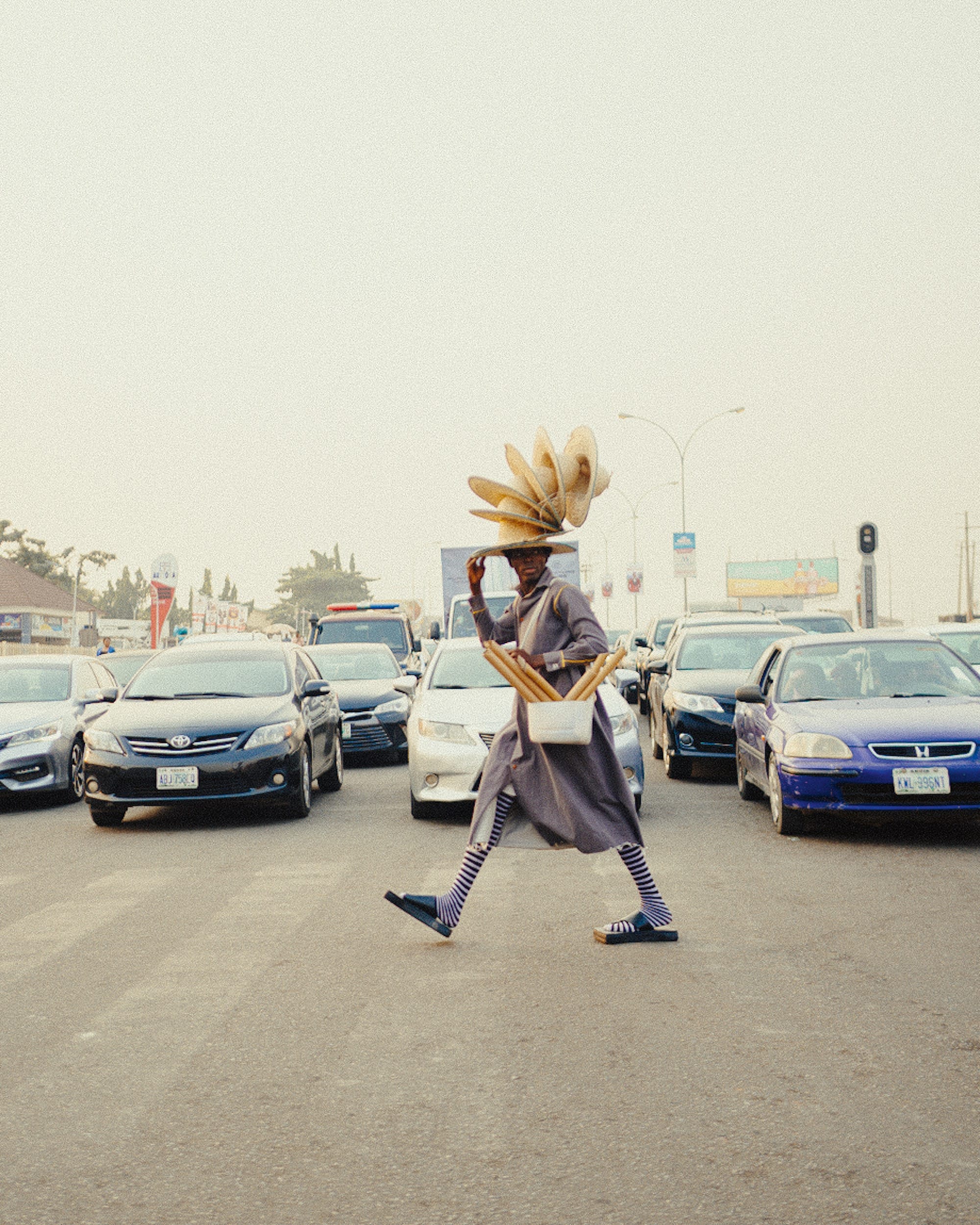 a man with numerous hats on his head crosses the street in front of cars