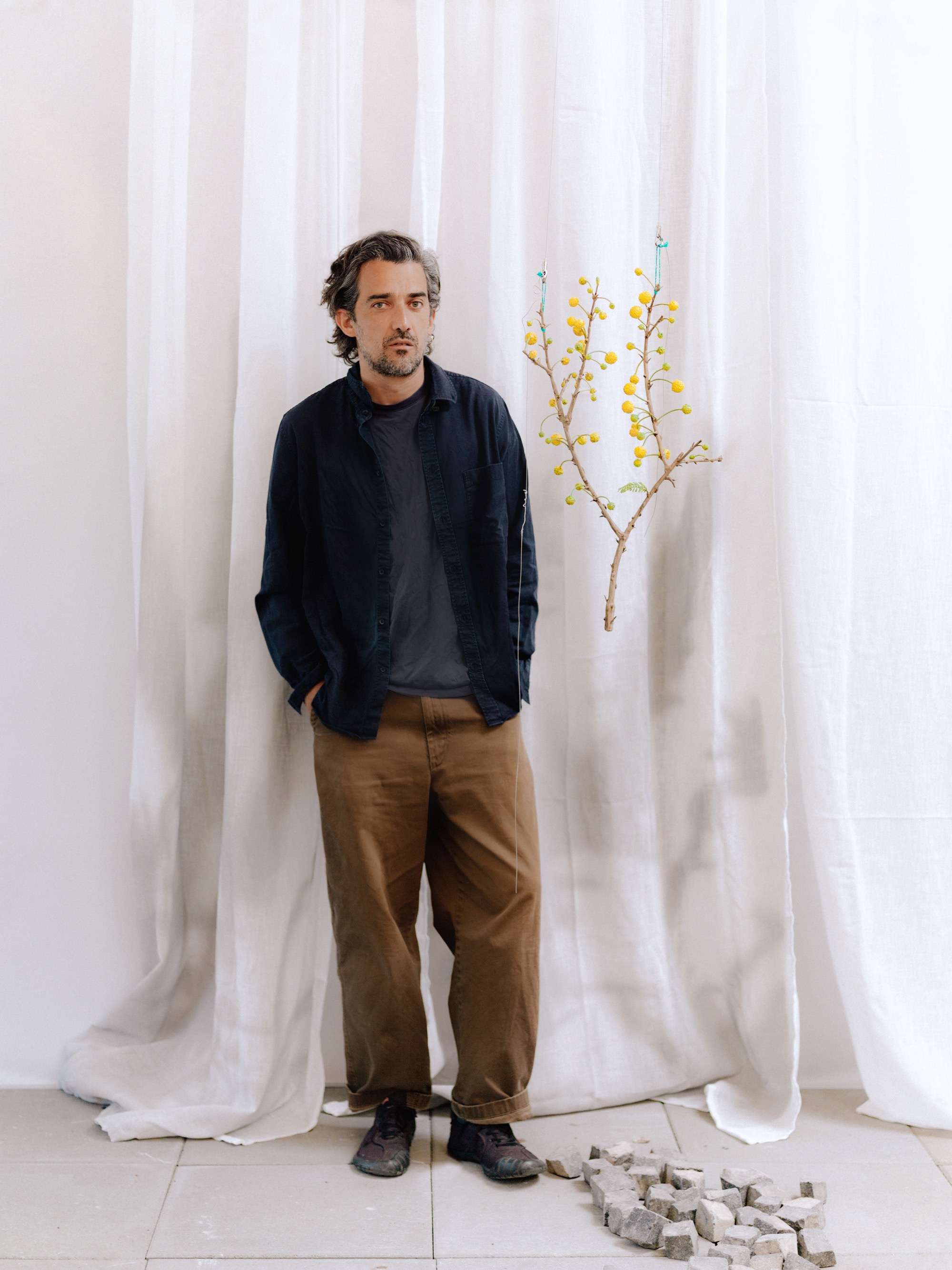 a man in a dark blue overshirt and undershirt and brown pants stands with his hands in his pockets next to a suspended botanical sculpture and a sheet